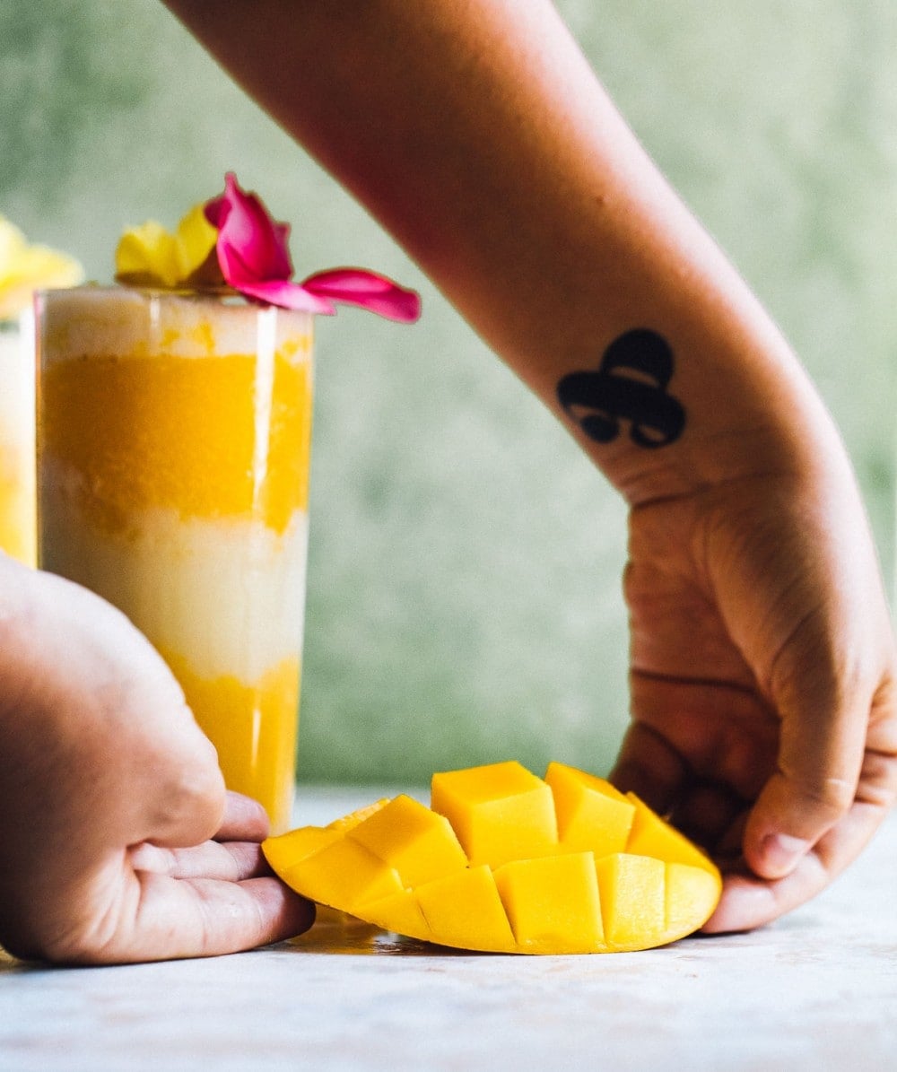 holding a mango that has been cut like a hedgehog with a cocktail in the backround