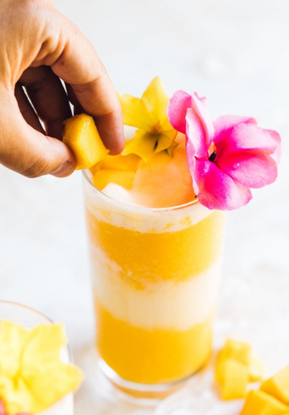 putting a mango cube onto a glass with a mango cocktail inside the glass. pink and yellow flowers are used as a garnish.