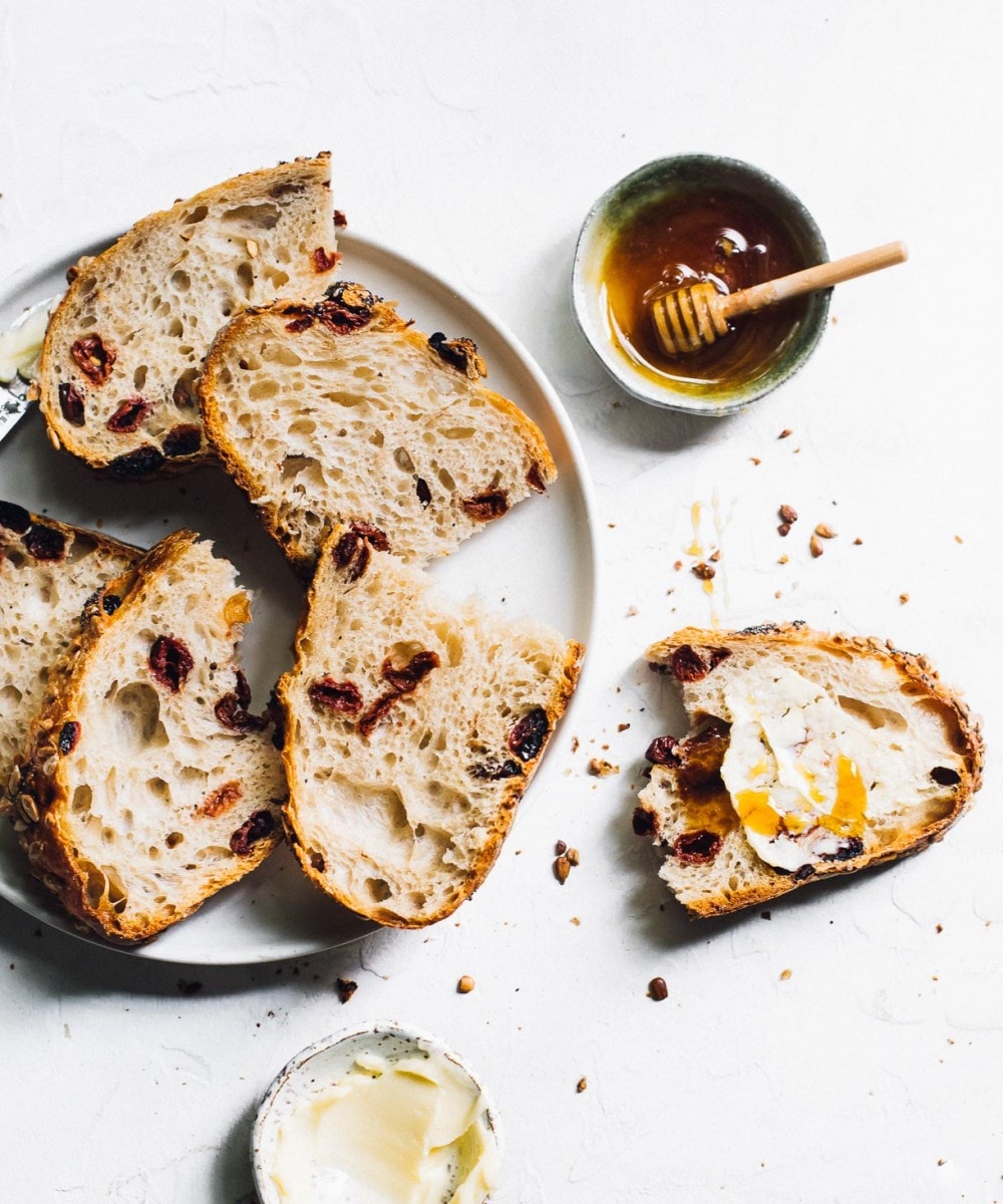 Tart Cherry Sourdough bread slices on a white plate, and one half eaten slice on the right of the plate slathered with butter and honey