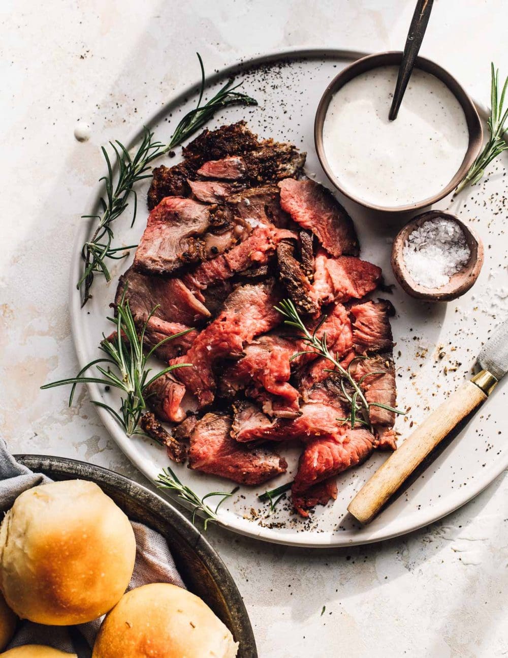 shaved prime rib on a white plate, with dinner rolls in bottom left