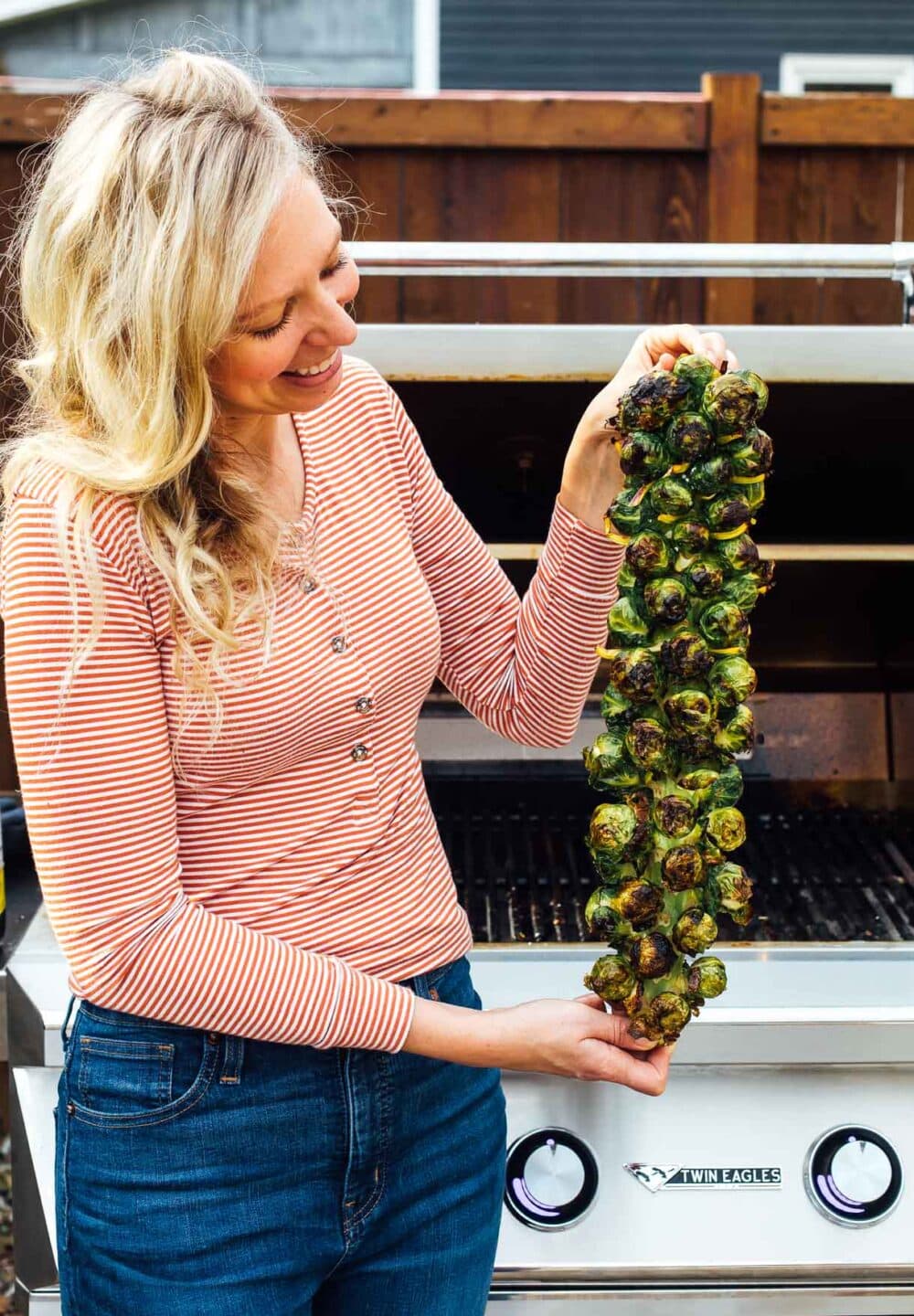 woman holding grilled brussels spouts on the stalk standing in front of a grill