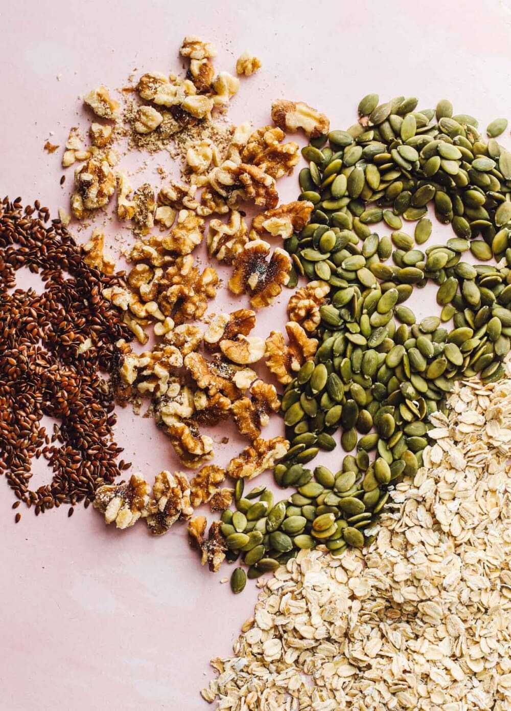flax seed, walnuts, pepita seeds, and oats laying on a pink surface
