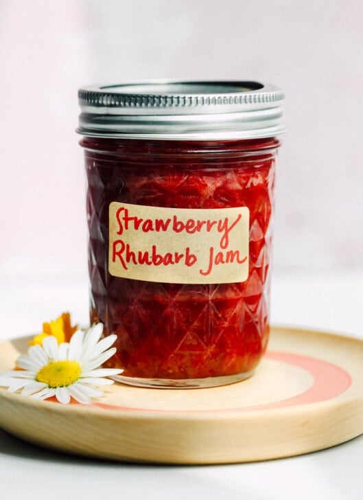 strawberry rhubarb jam in a glass jar with label, sitting on a wooden coaster