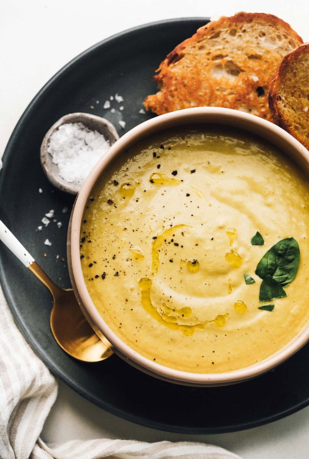 Bowl with green tomato soup on blue plate, with bowl of salt and sourdough bread.